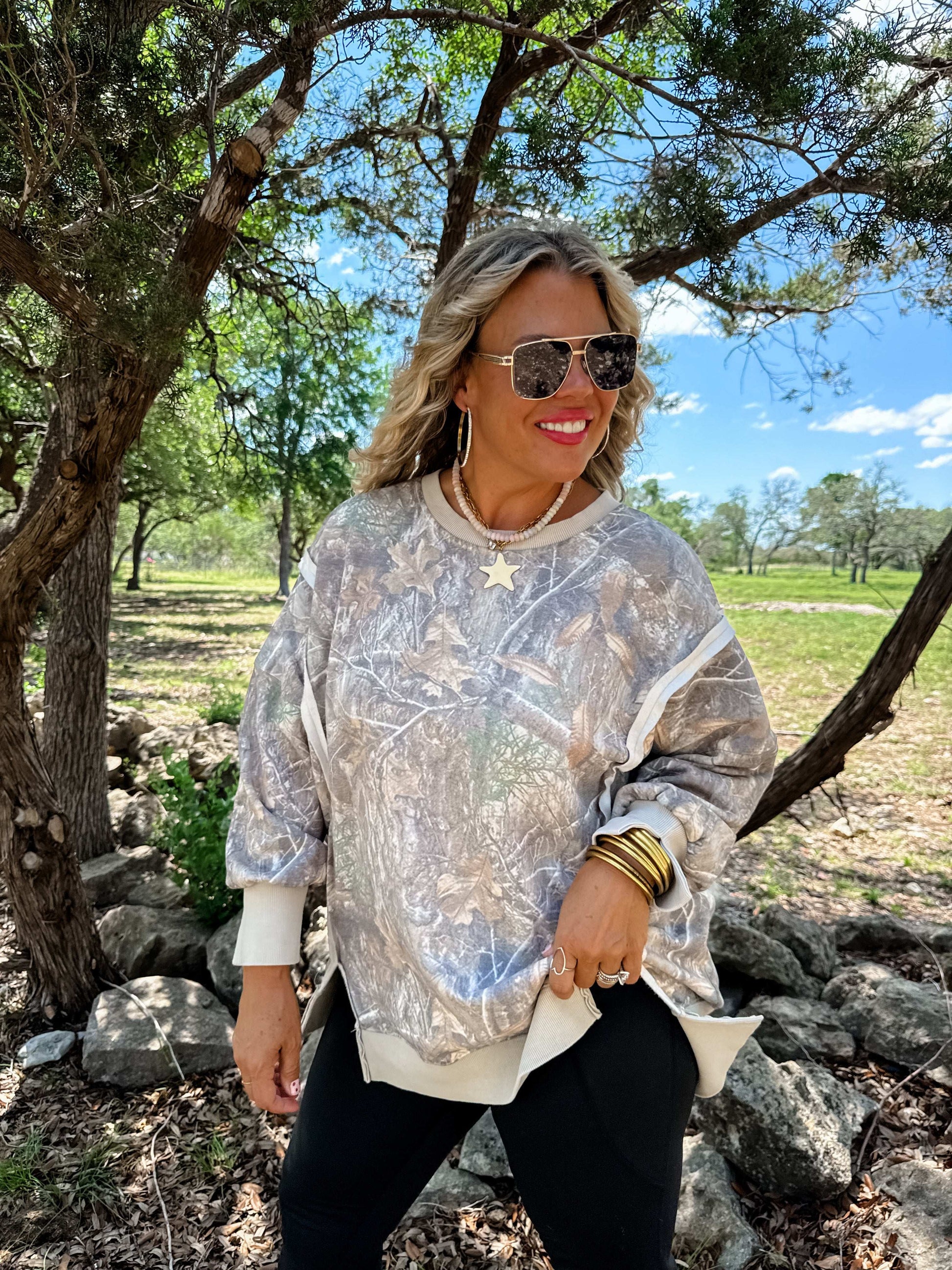 Woman wearing a camo top and sunglasses outdoors with trees and sky in the background