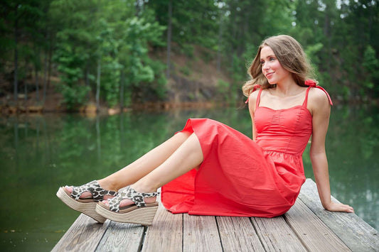 Woman in a red dress sitting on a wooden dock by a lake with trees in the background