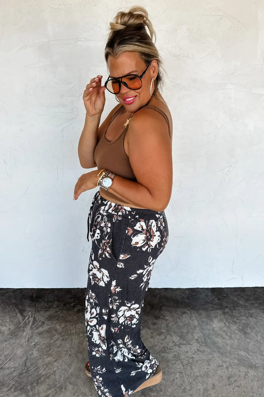 Woman wearing a brown top and floral pants against a white wall.
