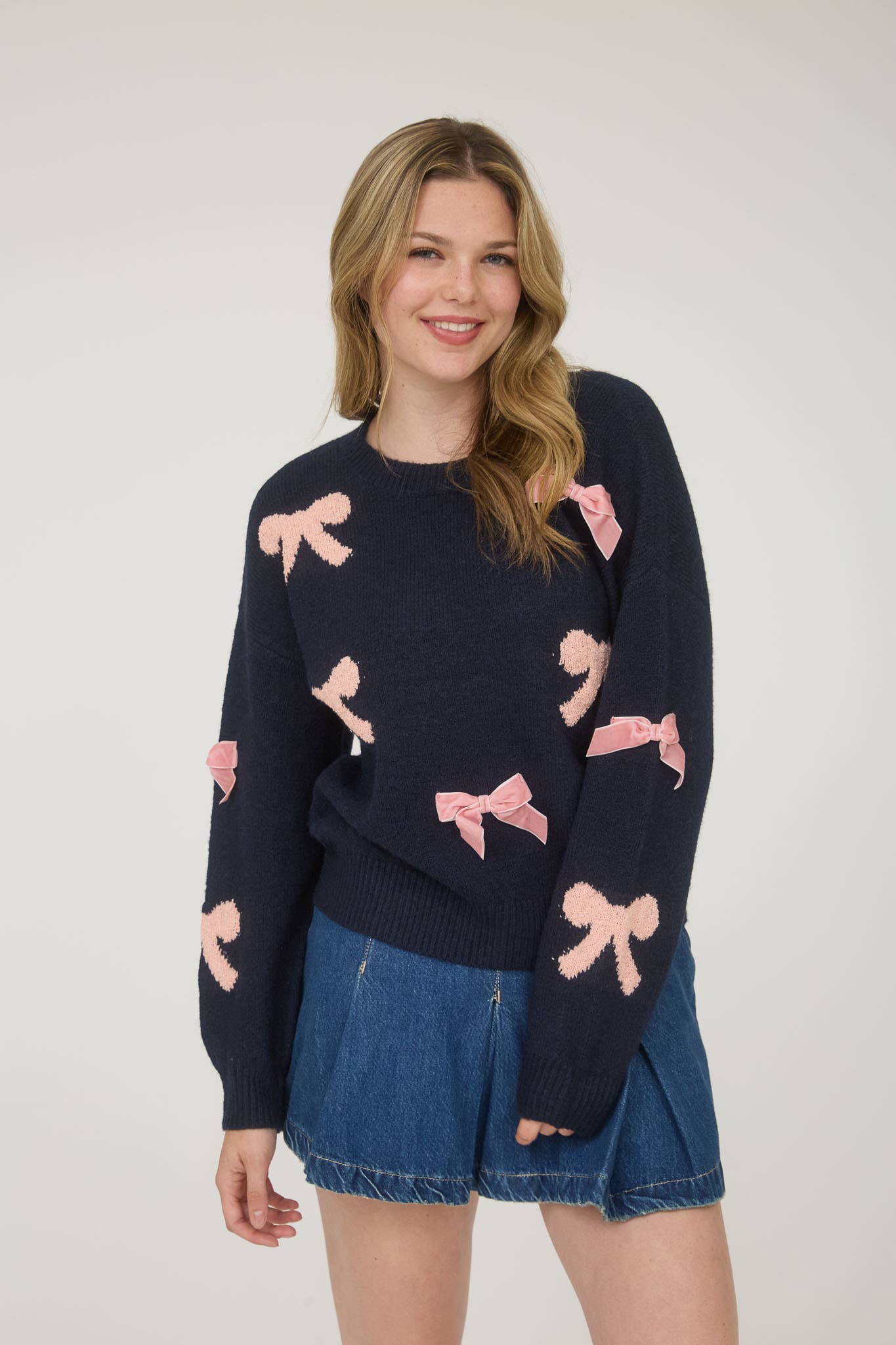 Woman wearing a navy sweater with pink bow patterns and denim shorts on a plain background