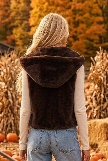 A woman shown from the back wearing a dark brown faux-fur hooded vest over a light beige long-sleeve top and blue jeans, against a solid dark gray studio background.