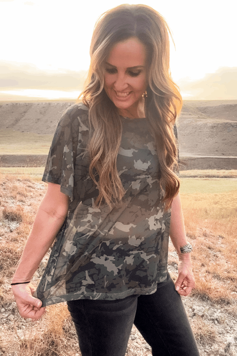 Woman wearing a camouflage shirt in an open field with a sunset.