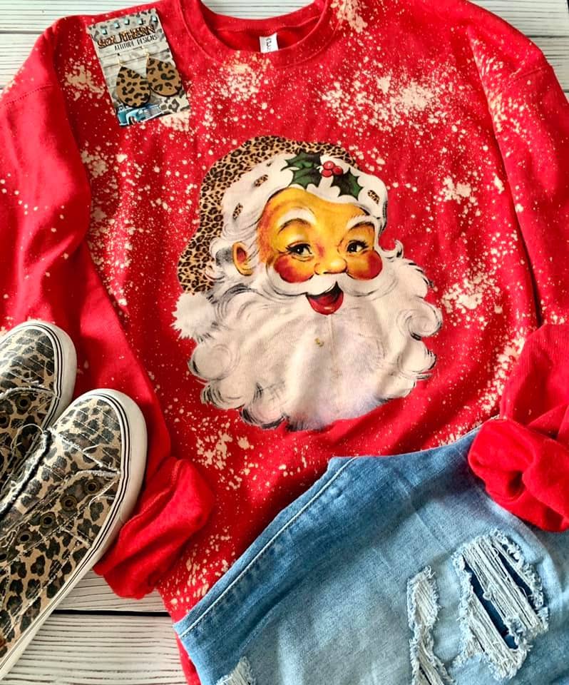 A red Christmas crewneck sweatshirt featuring a large Santa face graphic with leopard-print accents, styled in a flat lay with leopard-print sneakers, distressed denim shorts, and a leopard-print tag on a light wood background.