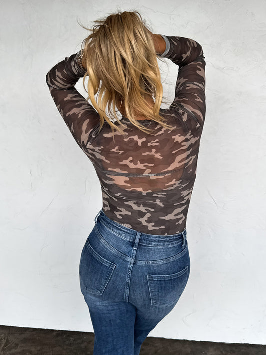 Person wearing a camouflage long-sleeve top and blue jeans against a white wall.