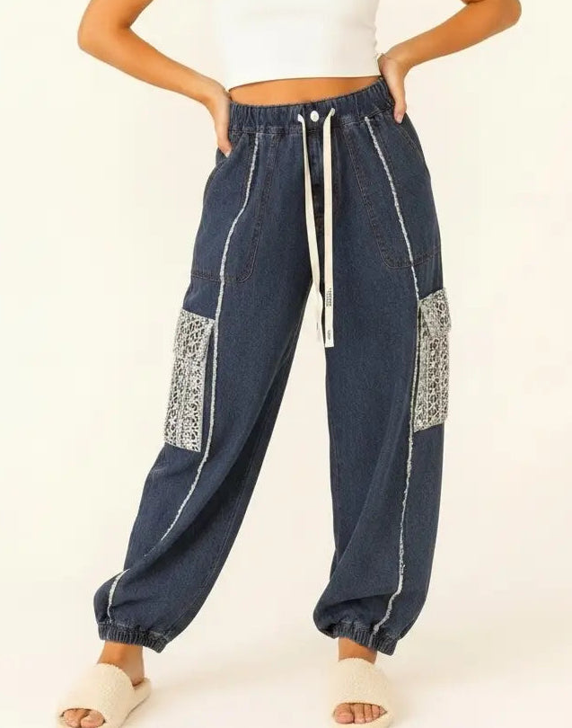 A woman modeling a pair of dark blue denim jogger pants with white contrast stitching, white drawstrings, and silver patterned patch pockets, paired with a white crop top and white slide sandals.