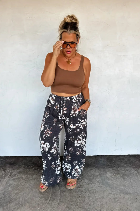 Woman wearing a brown crop top and black floral pants against a white wall.