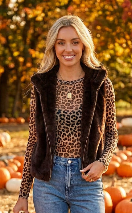 A woman modeling a dark brown faux fur vest with gold-tone zippers over a beige long-sleeve top and blue jeans, set against a solid dark gray studio background.